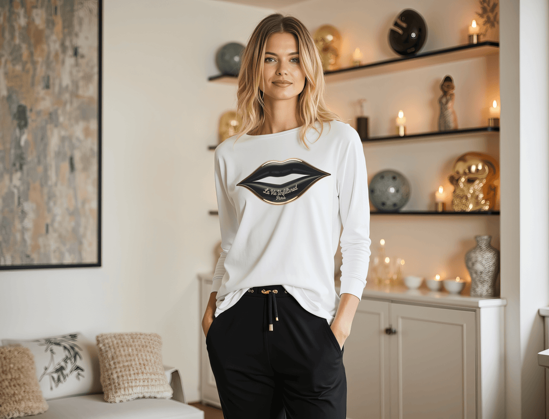 Woman wearing a white long sleeve top with a black lips graphic standing in a modern living room with shelves and lit candles