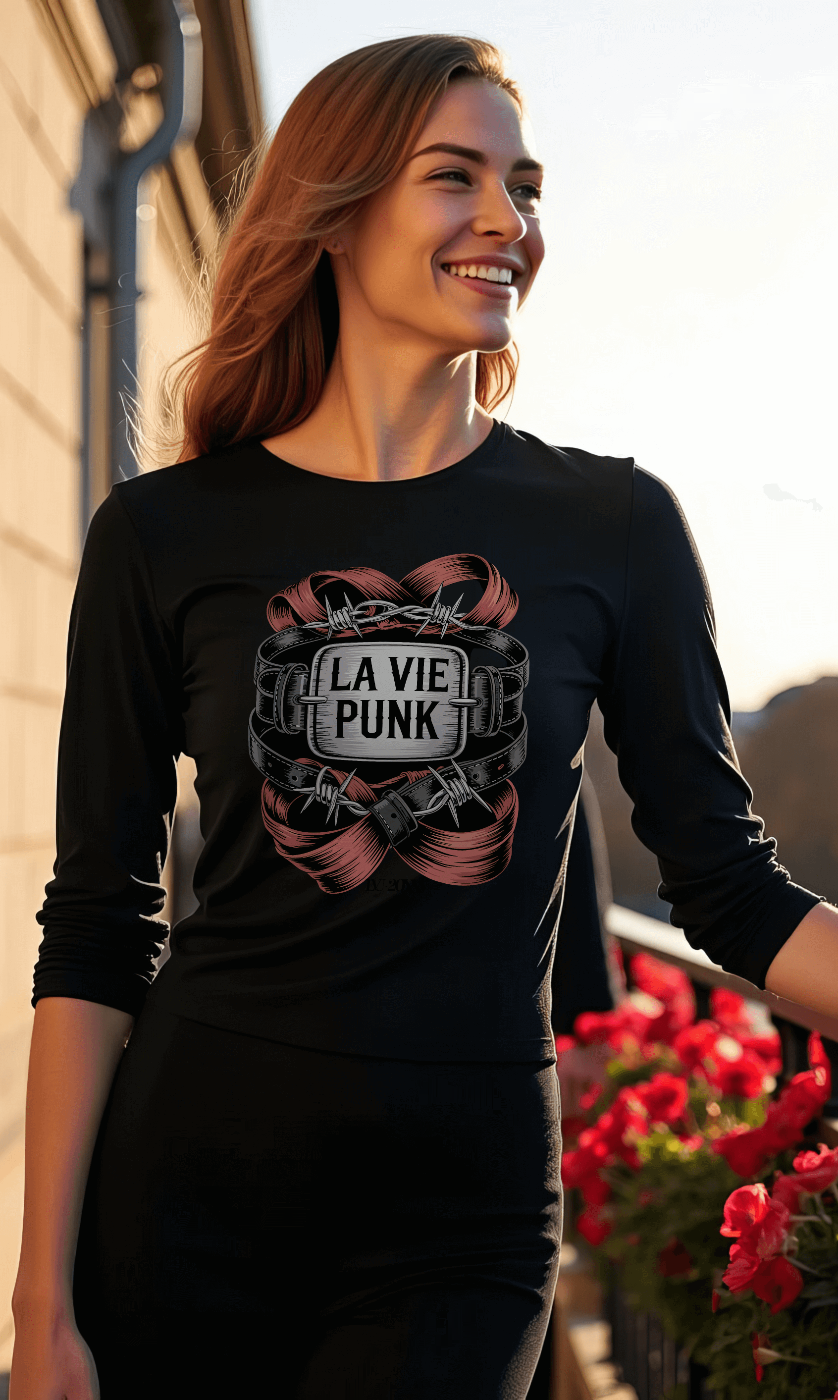Model smiling in Paris wearing a black long-sleeve La Vie Punk graphic top with ribbon and crest design, styled outdoors with flowers and architecture in the background.