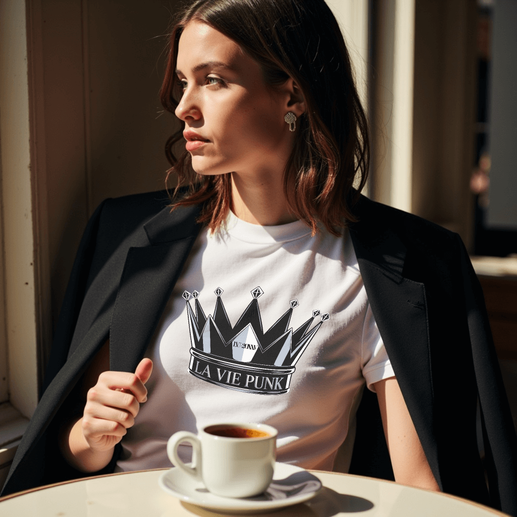 Model seated at a Parisian café wearing the Throne Punk Allure white jersey tee with crown graphic and 'La Vie Punk' lettering, styled with a black blazer, sipping coffee in natural morning light.