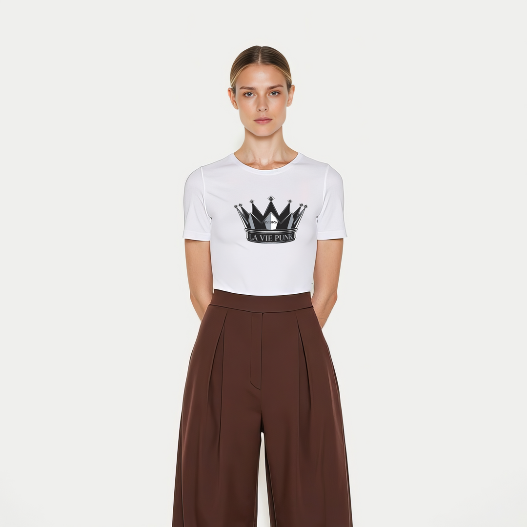 Model wearing the Throne Punk Allure jersey tee in white with a bold black crown graphic and 'La Vie Punk' lettering, styled with high-waisted brown wide-leg trousers, photographed on a plain white background.