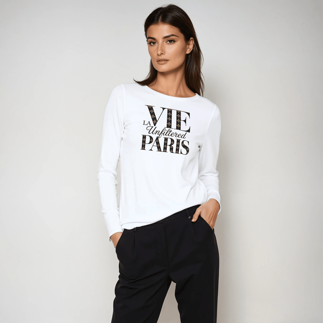 Model in white long-sleeve tee with bold 'La Vie Unfiltered Paris' graphic, styled with black tailored trousers in a studio setting.