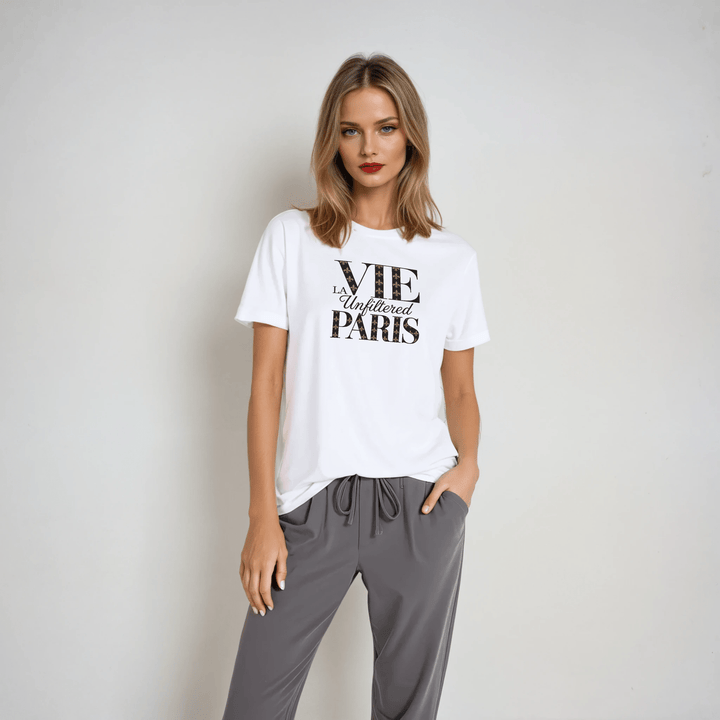 Model wearing white boxy-fit short sleeve tee with black 'La Vie Unfiltered Paris' graphic, paired with relaxed gray drawstring pants in a studio setting.