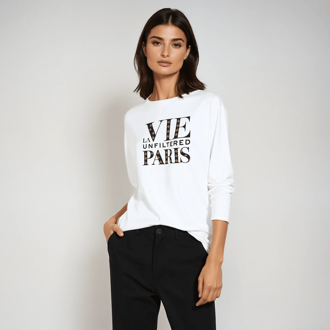 Model in white long-sleeve tee with bold 'La Vie Unfiltered Paris' monogram graphic, styled with black trousers in a studio setting.