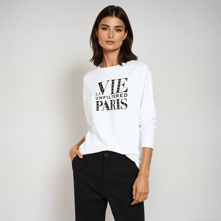 Model in white long-sleeve tee with bold 'La Vie Unfiltered Paris' monogram graphic, styled with black trousers in a studio setting.