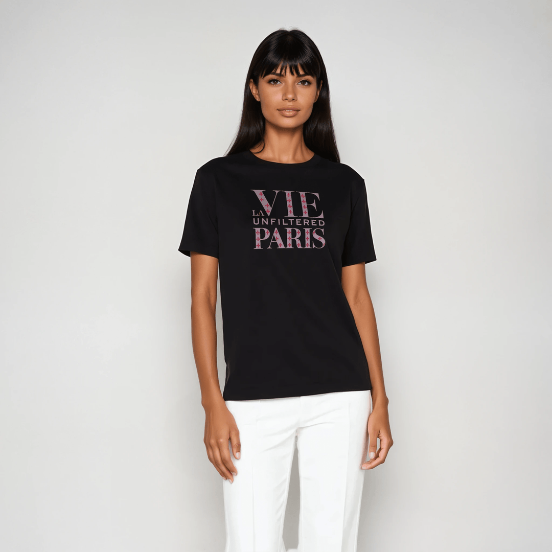 Model wearing black boxy-fit short sleeve tee with rose pink 'La Vie Unfiltered Paris' monogram graphic, styled with white tailored pants in a studio setting.